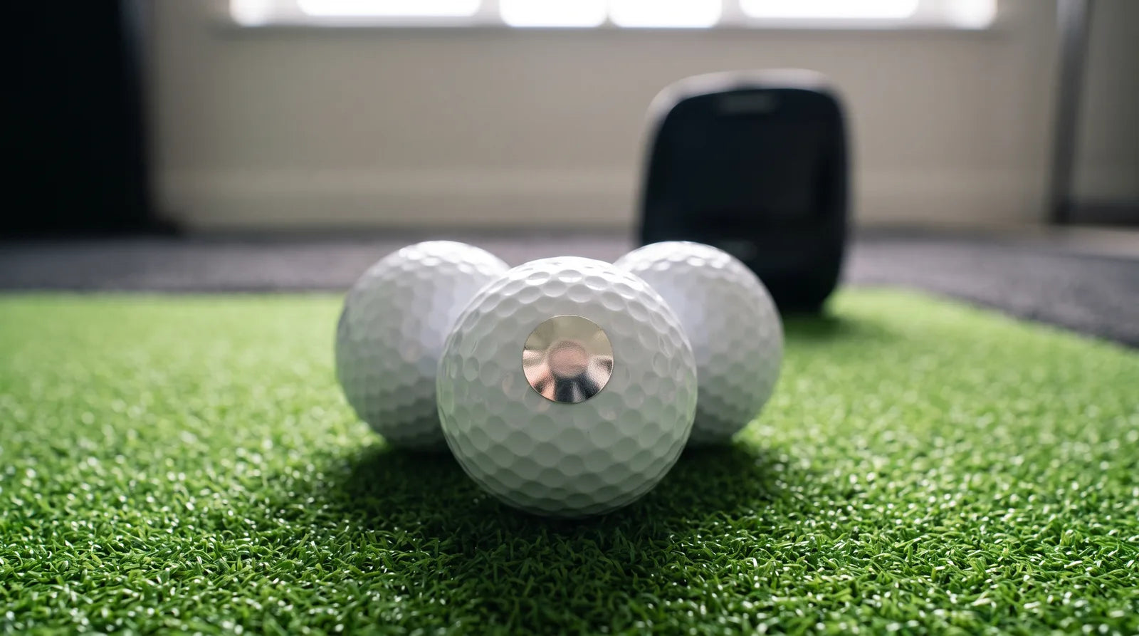 A macro photo of 3 white golf balls on green hitting turf, one with a small round metallic sticker on its cover, with a launch monitor out of focus behind